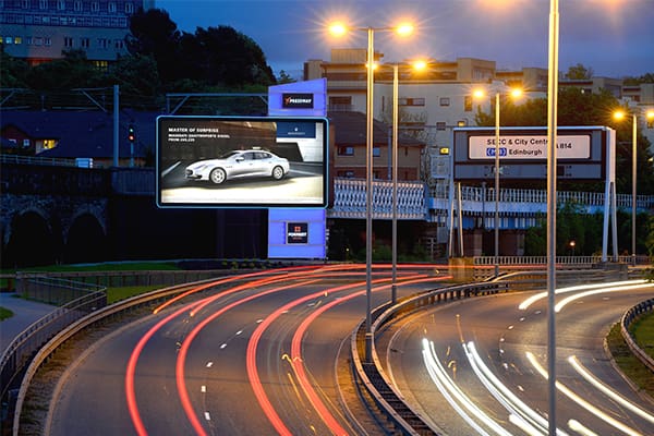 A busy multi-lane highway at dusk with light trails from moving vehicles. Illuminated billboards featuring LED backlit graphics line the road, one displaying an advertisement for a luxury car. Buildings and trees are visible in the background under a darkening sky, capturing the essence of out-of-home advertising.