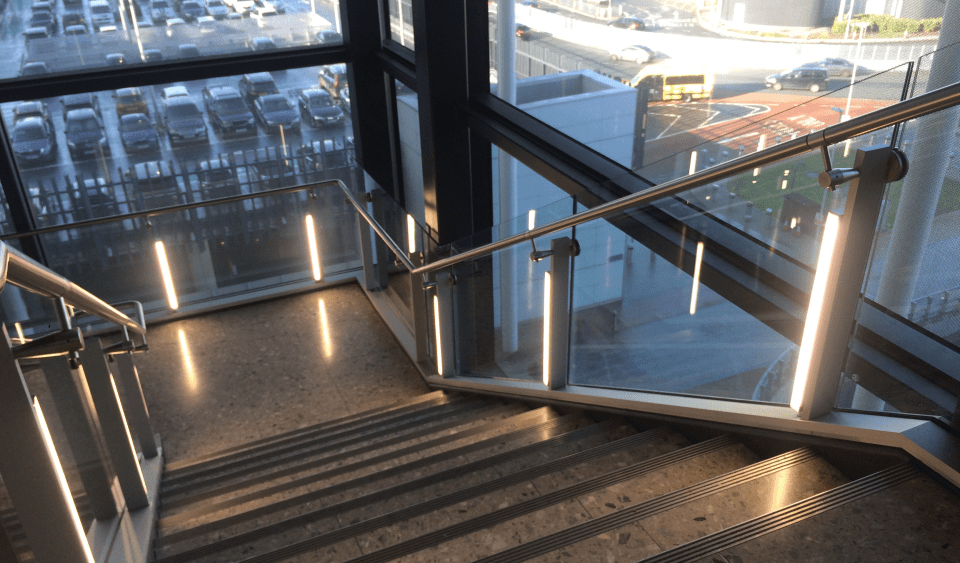 A modern stairwell with metal railings and LED illuminated handrails leads downwards. Large windows on the sides provide a view of a sunlit parking lot and city street outside. The stairwell is well-lit with natural light streaming in.