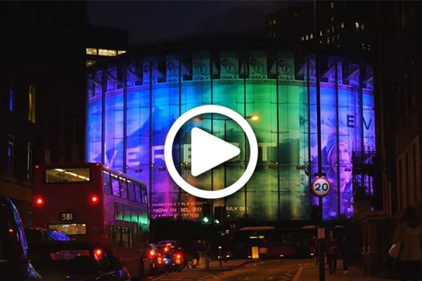 A large, cylindrical building illuminated with a colour changing LED lighting system, casting vibrant blue, green, and purple hues at night. A double-decker bus and several cars are on the street in front of the IMAX-like structure. A play button icon is centered over the image.