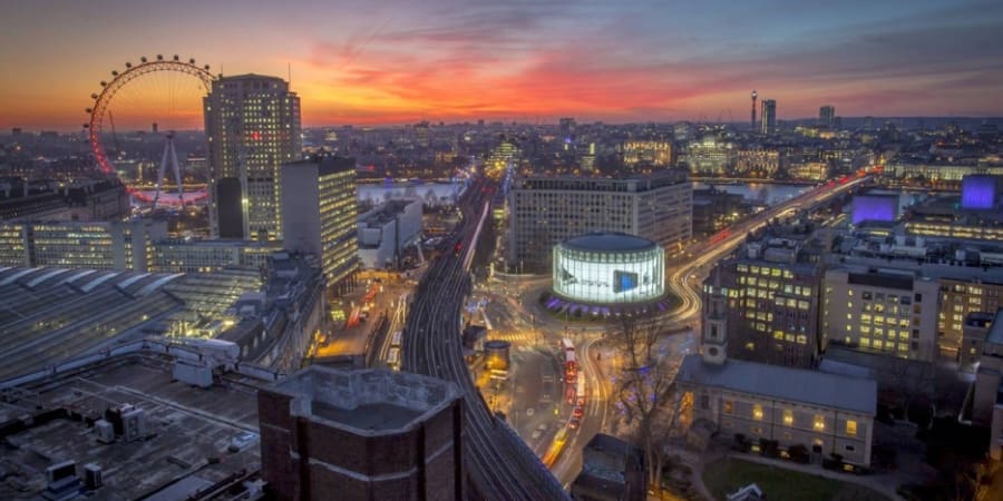 A cityscape view of London at sunset. The London Eye is visible on the left side, with the city's lights beginning to illuminate the landscape. Nearby, an IMAX illuminated by Bright Green Technology adds a modern touch. Buildings and roads are bathed in the warm glow of the setting sun, creating a vibrant and dynamic urban scene.