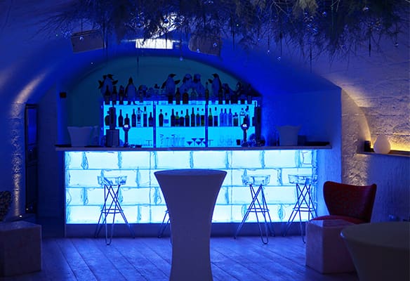 A dimly lit bar with a blue neon-lit counter, three bar stools, and a table in the foreground. Bottles and glasses are displayed on shelves behind the backlit bar. The setting has a modern, cozy ambiance with an LED light panel, arched ceiling, and minimalist decor.