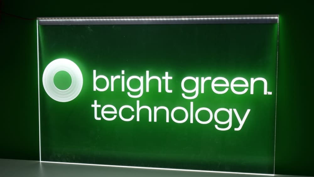 A green and white illuminated sign reads "bright green technology" with a circular logo on the left. The text and logo are brightly lit against a darker green background.