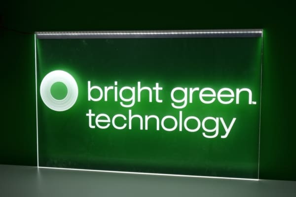 A green and white illuminated sign reads "bright green technology" with a circular logo on the left. The text and logo are brightly lit against a darker green background.