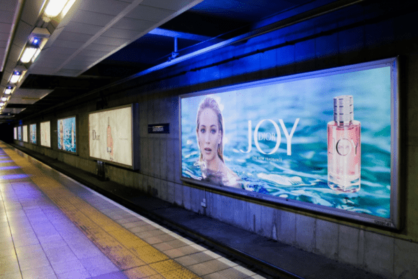 A subway platform features several large, LED backlit graphics for Dior. The brightly lit ads showcase Dior's "JOY" perfume, with an image of a woman submerged in water next to a bottle of the fragrance. This out of home display illuminates the platform, which is lined with lights and has a tiled floor.