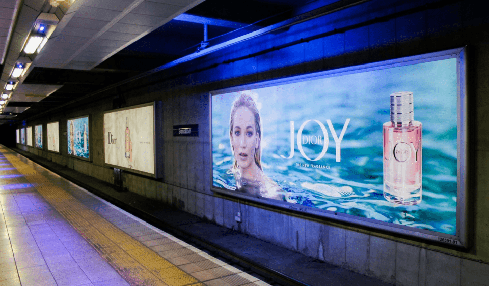 A subway platform features several large, LED backlit graphics for Dior. The brightly lit ads showcase Dior's "JOY" perfume, with an image of a woman submerged in water next to a bottle of the fragrance. This out of home display illuminates the platform, which is lined with lights and has a tiled floor.