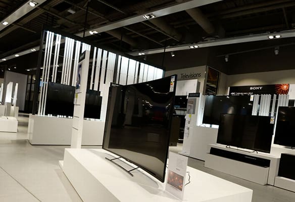 A modern electronics store with various television sets on display features sleek, minimalist design elements and ample lighting. Several TVs are placed on white stands next to illuminated acrylic displays, with signage and branding visible in the background.