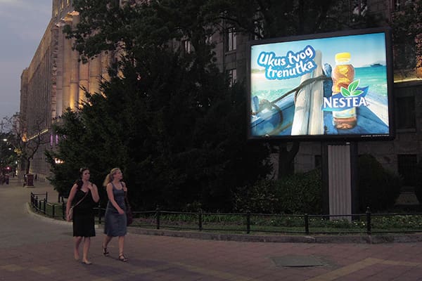 Two people walk on a sidewalk in front of a large illuminated billboard featuring LED backlit graphics. The advertisement is for NESTEA, displaying a tropical-themed image and the text "Ukus tvog trenutka." The scene is set during dusk with dim natural and artificial lighting, highlighting this out of home advertising.