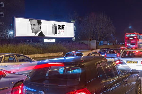 Cars are stuck in traffic at night under a large LED backlit billboard featuring a male model and an advertisement for Mont Blanc. This urban out of home scene captures the bustling city life, with a bus visible on the road to the right.