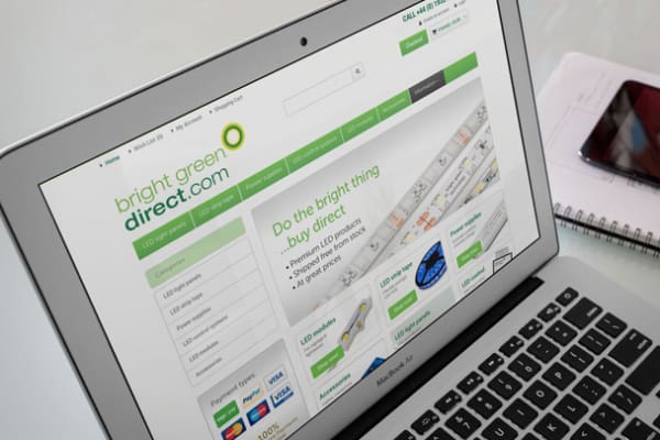 A laptop screen displays the homepage of "bright green direct.com," featuring various categories and products related to LED lighting. Next to the laptop, there is a notepad with a pen and a smartphone resting on top of it.