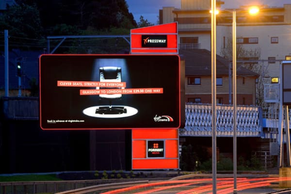 A digital billboard for Virgin Trains, with vibrant LED lights, displays an advertisement promoting travel from Glasgow to London with fares starting at £29.50 one way. The sign reads: "Clever seats. Strictly for everyone!" This out of home ad shines brightly on an urban street during the evening.