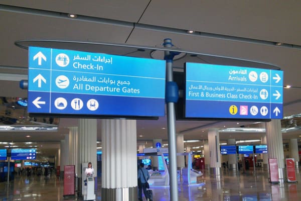 A modern airport terminal features blue LED wayfinder signage directing travelers to check-in, all departure gates, arrivals, and first & business class check-in. The signs also include icons for baggage, restrooms, and other amenities. The terminal is spacious and well-lit.