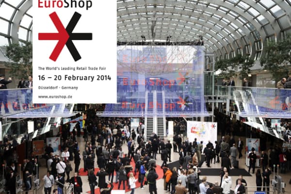 EuroShop 2014 trade fair in Düsseldorf, Germany. Crowds of attendees walk and gather in a large, modern hall with a curved glass roof. EuroShop logo and event details are displayed on a banner. Exhibition stands and information booths are visible in the background.