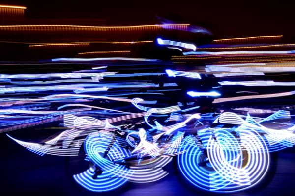 A cyclist rides through a dark scene with a colour changing LED lighting system on the bike, creating vibrant light trails that blend together in motion. The blurred effect captures dynamic movement and colorful patterns against a dark background.