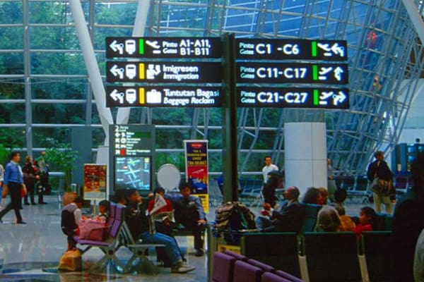 A busy airport terminal with travelers seated in waiting areas. LED wayfinders display directions to various gates and services, including immigration and baggage claim. The terminal features large windows and a high ceiling with a modern architectural design.