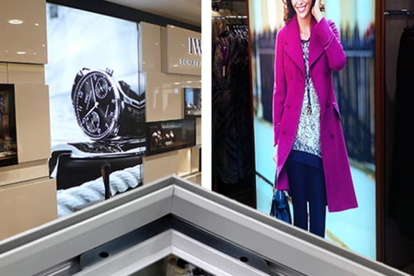 A retail store display featuring a large image of a wristwatch and another of a woman wearing a vibrant pink coat, both presented as backlit graphics. The images are part of the store's promotional material, and there is an LED lightbox in the foreground showcasing highlighted products.