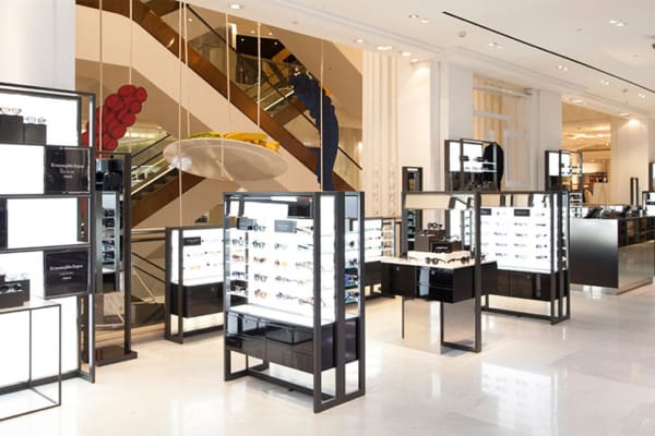 A modern and sleek eyewear store with multiple illuminated display cases showcasing various pairs of glasses. The store features clean, white walls and glossy floors, enhanced by LED light sheet panels, with a staircase and escalator in the background. The ambiance is bright and inviting.