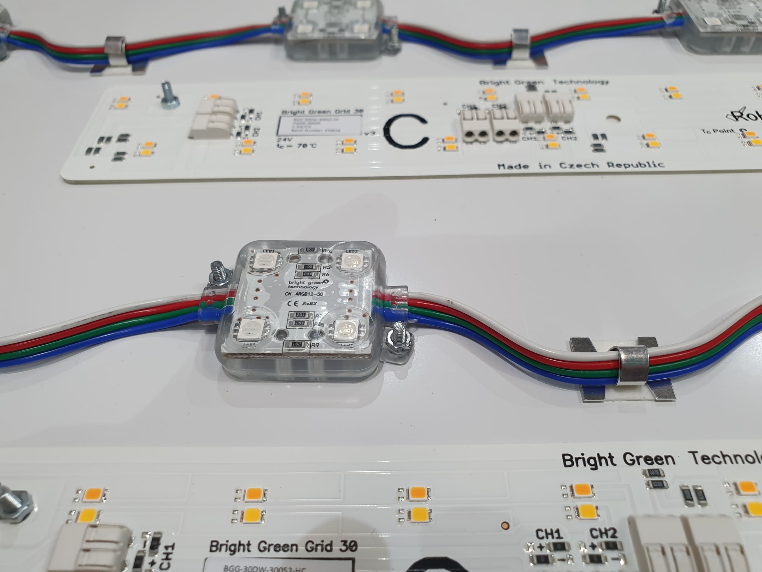 Close-up of various LED strip lights and small LED modules wired together on a white surface. The LED modules have multicolored wires attached, and the strips display small yellow LED bulbs along with some text and symbols printed on them.