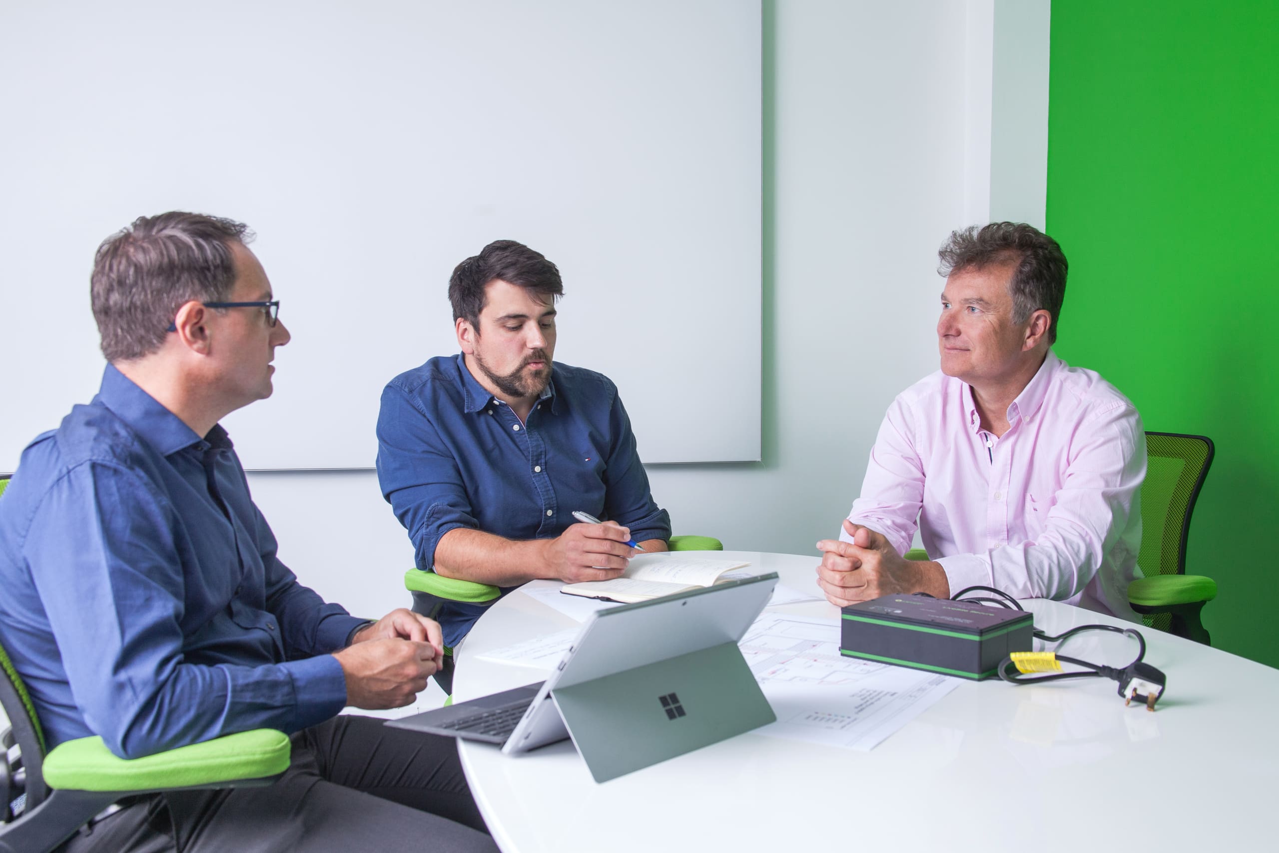 Three men sit around a white table in a modern office, engaged in a discussion. Two men are on the left, one in a blue shirt taking notes, the other in a dark blue shirt talking about an LED driver project. The third man, in a light pink shirt, listens attentively. A tablet and paperwork are on the table.