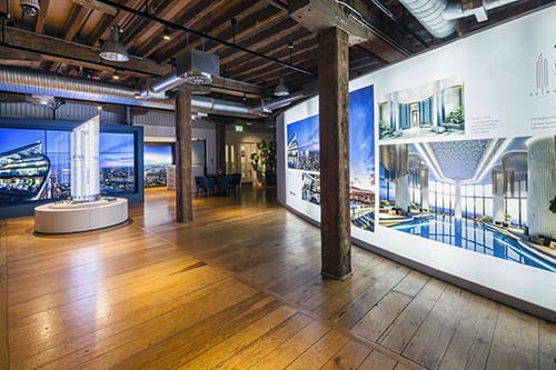 A modern exhibition space with wooden floors and exposed wooden beams. Large LED backlit graphics display architectural renderings and photographs of sleek, contemporary buildings. A detailed scale model of a high-rise structure is centrally placed.