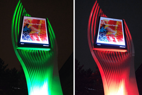 A modern, white, curved digital billboard is shown at night. The left side of the image displays the LED-lit billboard glowing with green and white lights like a futuristic advertising sculpture. The right side features red and green lights, showcasing a vibrant advertisement.