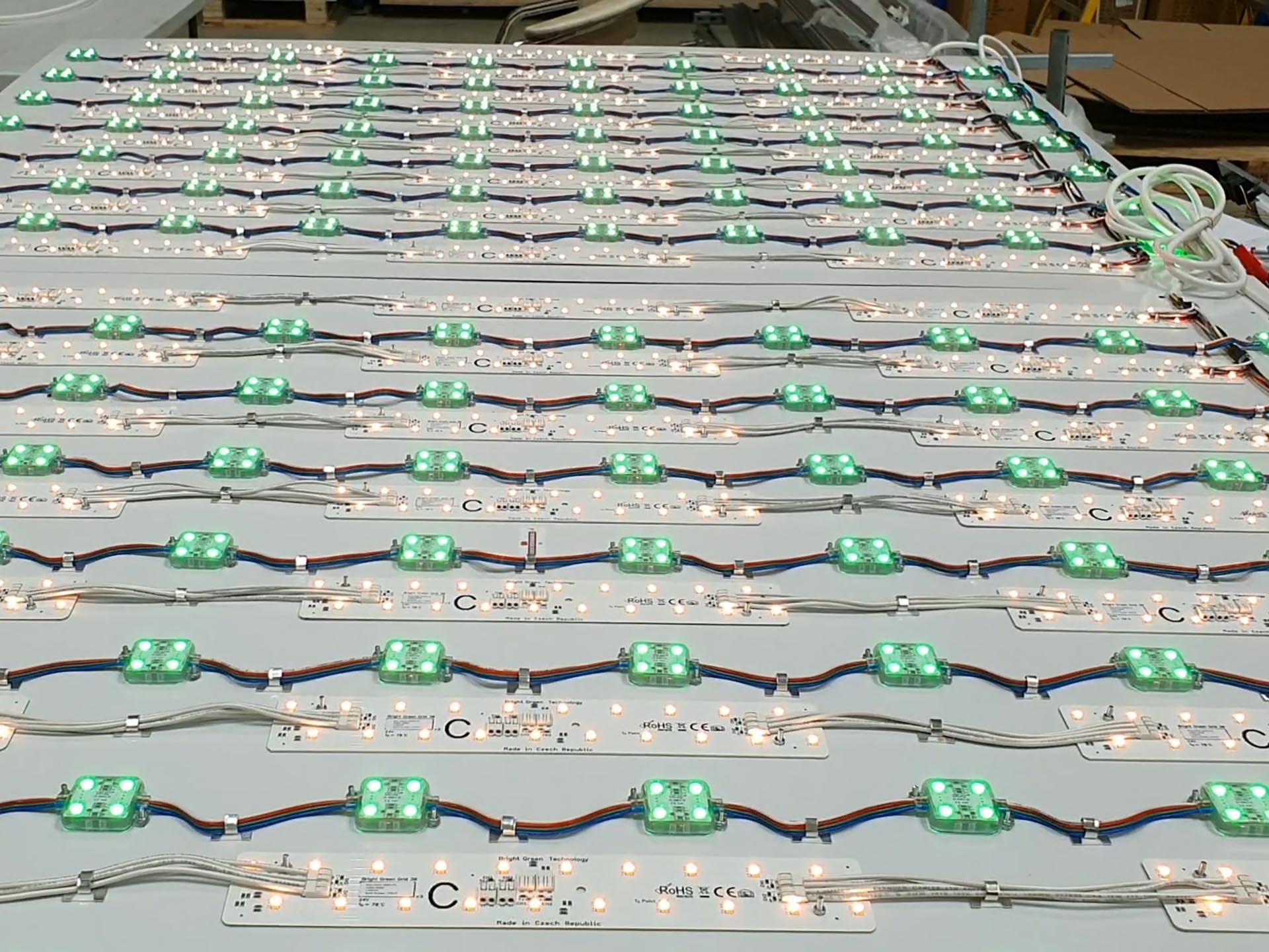 A large panel features an array of green LED lights and electrical wiring. Each LED unit is connected with multicolored cables in a precise symmetrical grid pattern. The background has various electronic components and tools, indicating a workshop or assembly area.