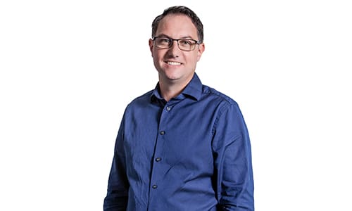 A person with short dark hair, glasses, and a light smile is wearing a blue button-up shirt. They are standing against a plain white background.