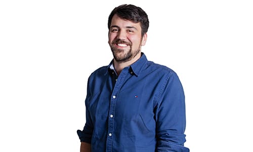 A man with a beard and short dark hair is smiling at the camera. He is wearing a blue button-up shirt with the sleeves rolled up. The background is white.