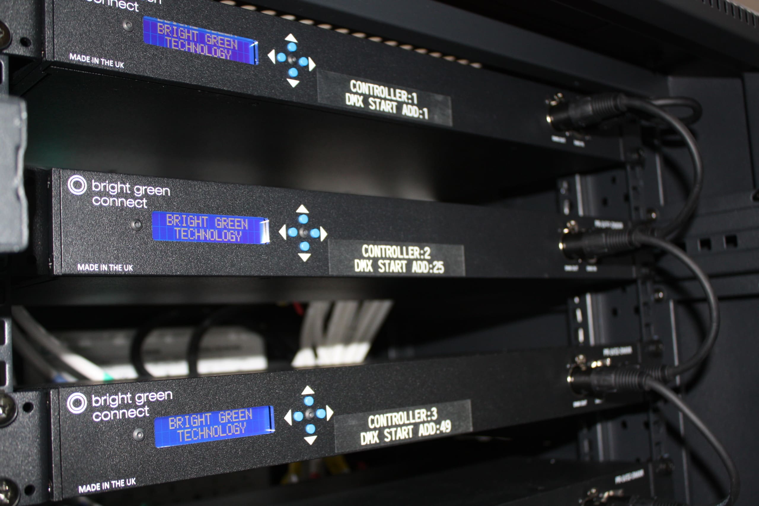 Three black electronic devices with blue LED displays, boasting Flicker Free LED Lighting, are mounted in a server rack. The displays read "BRIGHT GREEN TECHNOLOGY," "CONTROLLER:1 DMX:START ADD:1," and other similar messages. Cables are connected to the devices, which bear the logo "bright green connect.