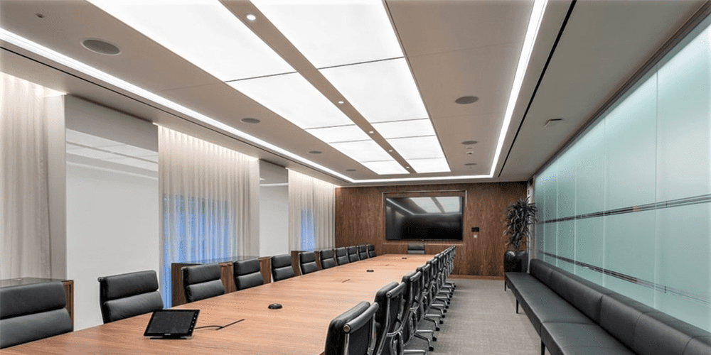 A spacious conference room at Carlyle Asset Management in London features a long, wooden table surrounded by black leather chairs. A large screen at one end is accentuated by wooden paneling, while the opposite wall showcases frosted glass and a long bench. The ceiling boasts custom-built modular framed systems with LED Light Panels and DALI dimmable drivers.