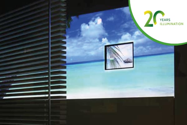 A window with horizontal blinds partially open reveals a sunny beach scene with blue water and a small island in the background. A small framed image of a palm tree leaf sits adjacent to an LED lightbox displaying a green and white logo that reads "20 Years Illumination.