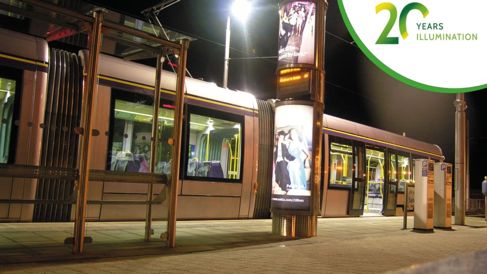 A modern tram at a lit-up station at night features the world’s first volume roll-out of LED backlit 6-sheet advertising panels on a bright vertical pillar. The station is empty with no passengers. In the top right corner, there's a logo for "20 Years of Illumination" in green and yellow.