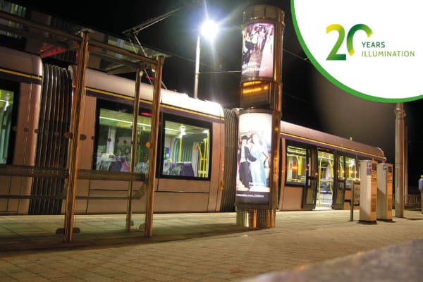 A modern tram at a lit-up station at night features the world’s first volume roll-out of LED backlit 6-sheet advertising panels on a bright vertical pillar. The station is empty with no passengers. In the top right corner, there's a logo for "20 Years of Illumination" in green and yellow.