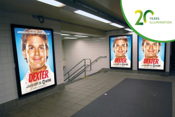 The New York City Underground subway station boasts tiled walls adorned with LED backlit posters for the TV show "Dexter." Each poster showcases a man smiling, his face marked with blood splatters, and a "20 Years Illumination" logo in the top right corner.