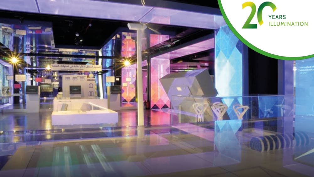 A modern museum exhibition space in Saudi Arabia with colorful LED illuminated showcases and interactive displays. The interior features various technological exhibits and large screens. A banner in the top right corner reads "20 Years Illumination" with a green graphic.