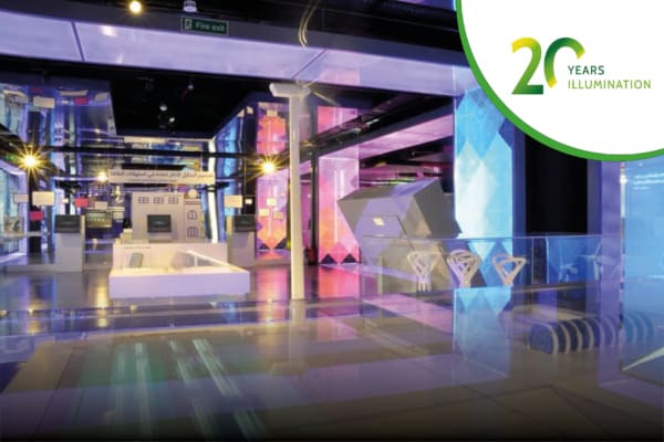 A modern museum exhibition space in Saudi Arabia with colorful LED illuminated showcases and interactive displays. The interior features various technological exhibits and large screens. A banner in the top right corner reads "20 Years Illumination" with a green graphic.