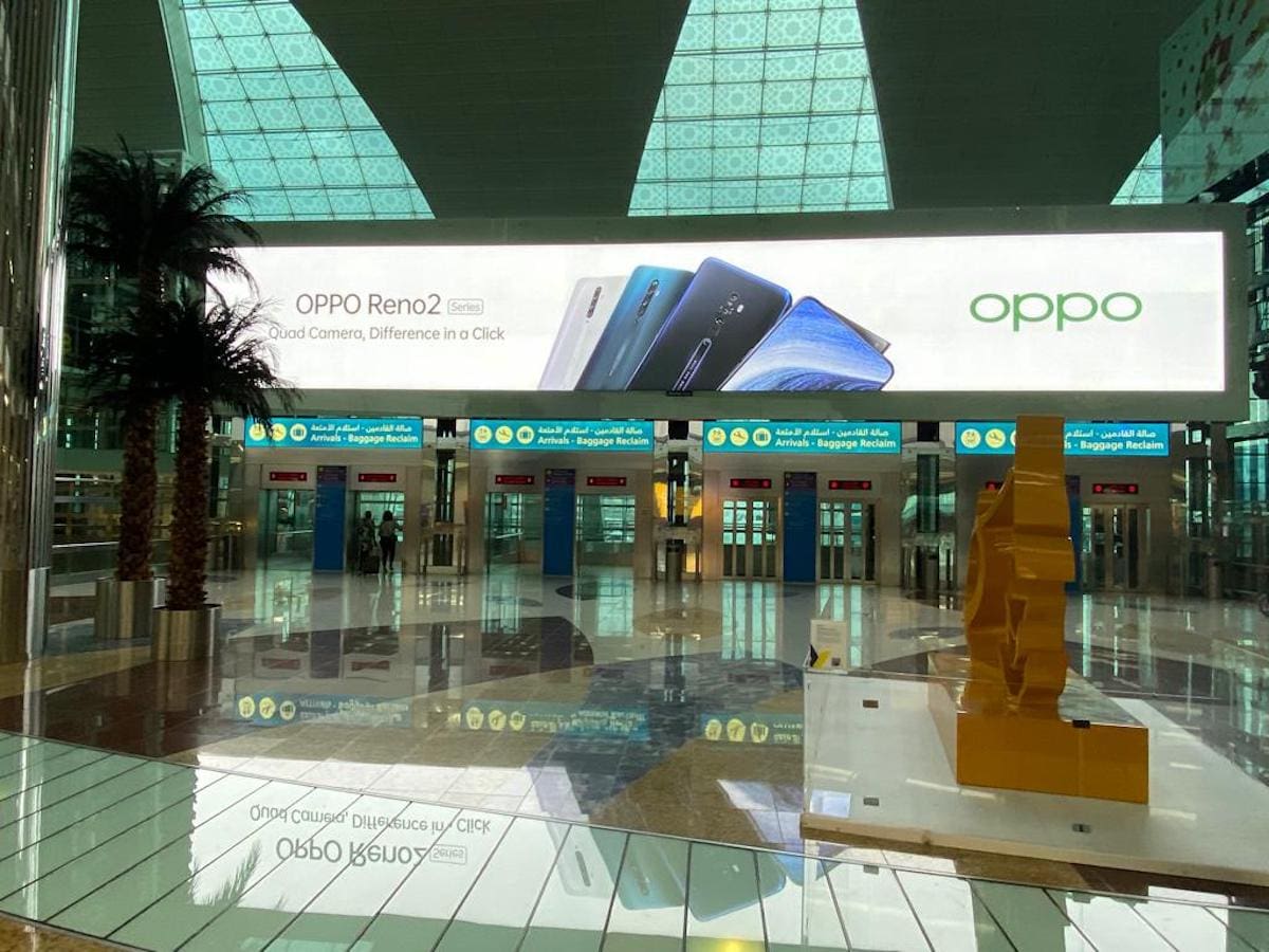 A spacious airport terminal with tall glass windows and a high ceiling. There is a large backlit advertisement hanging above the check-in counters, promoting the Oppo Reno2 smartphone with the tagline "Quad Camera, Diffidence in a Click." Multiple entrances and exits are visible below the advertisement.