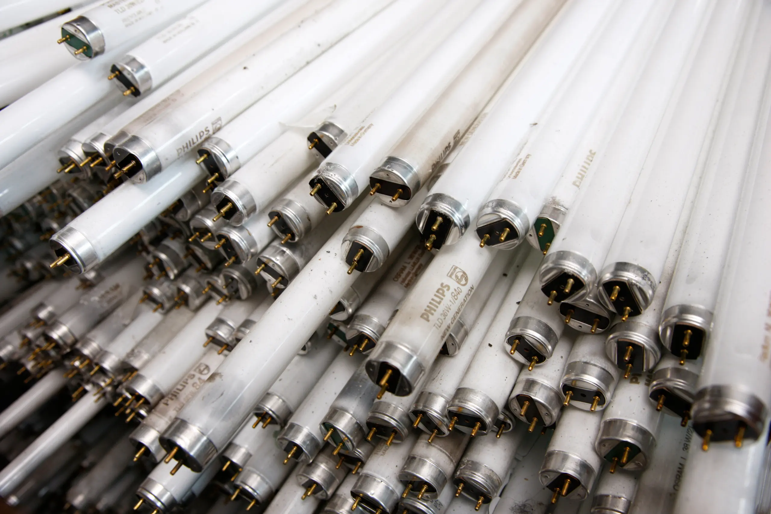 A large pile of used fluorescent light tubes, with metal caps and pins facing outward. The tubes are slightly dirty and show signs of wear. The photo suggests recycling or disposal of the bulbs, potentially making way for more efficient LEDs.