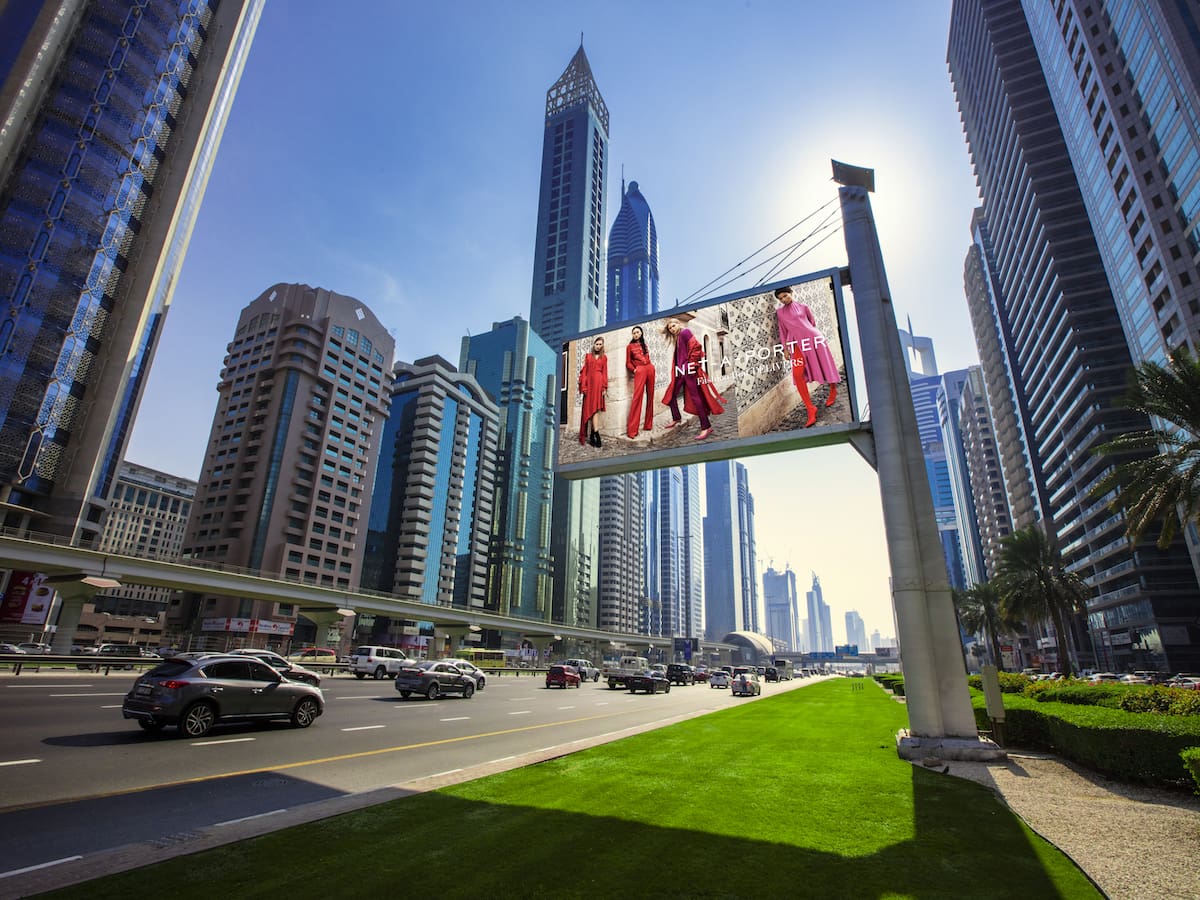 A vibrant urban scene showcasing towering skyscrapers under a clear blue sky. A digital billboard with backlit graphics features models in colorful clothing. Vehicles are driving on the road below, while palm trees and a well-maintained grassy area add to the foreground's charm.