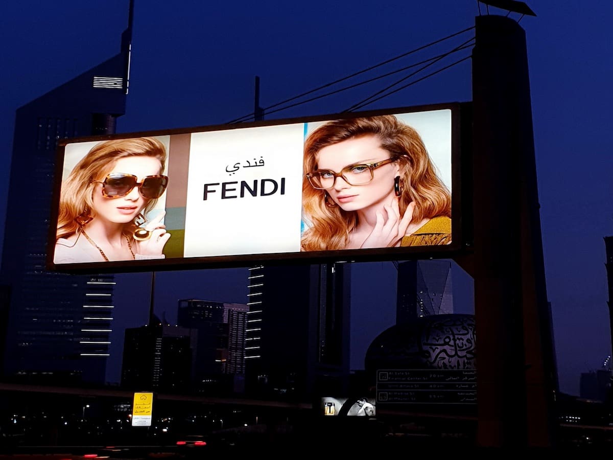 A backlit billboard featuring Fendi eyewear against a cityscape backdrop at dusk. The left side displays a woman wearing sunglasses, while the right side shows another woman in glasses. The Fendi logo is centered between the two images with some text in Arabic above it.