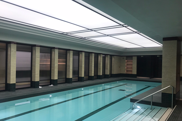 Indoor swimming pool with clear blue water, surrounded by beige walls and dark accents. Overhead, a bright, spacious ceiling with a modern design. Backlit surfaces enhance the ambiance as steps lead into the pool, and windows line one side of the room.