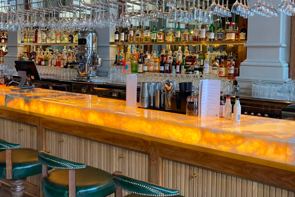 A luxurious bar featuring a glowing marble countertop and backlit surfaces, lined with green barstools. The shelves behind are stocked with various bottles of liquor and rows of hanging glasses, enhancing the elegant ambiance.