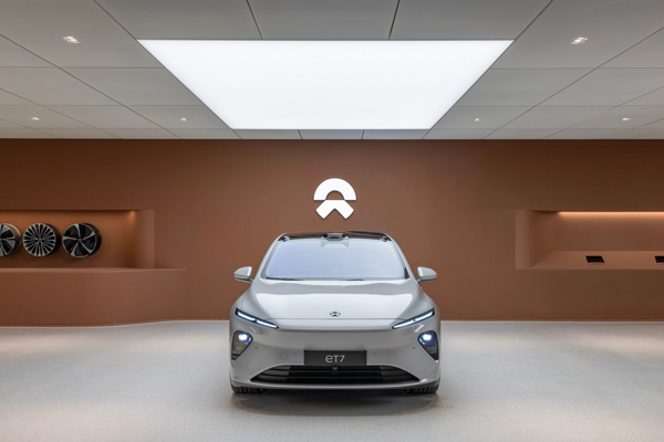 A sleek gray electric car, the NIO ET7, is showcased in a modern showroom with backlit surfaces and a minimalist design. A large ceiling light illuminates the space, highlighting the brown backdrop with the NIO logo and wheels mounted on the left wall.