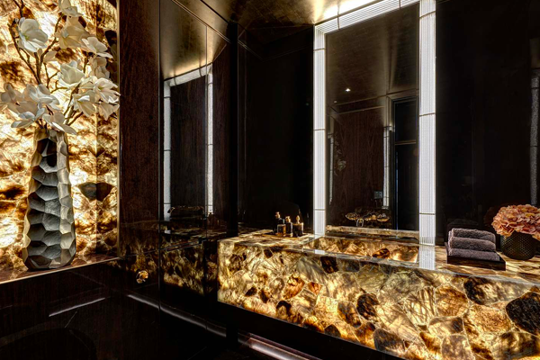 Luxurious bathroom with a glowing onyx vanity and backlit surfaces. Dark walls enhance the ambient lighting, while a sculpted vase with white flowers and folded towels are artfully placed, adding to the elegant atmosphere.